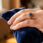 Gros plan sur la main d’une femme portant une ancienne bague en or ornée d’un saphir solidement serti, près d’une pochette en velours.
