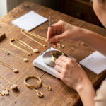 Une femme examine plusieurs bijoux en or sur une table en bois, avec une balance de précision, une loupe et des fiches d’estimation vierges, éclairés par une lumière naturelle.