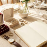 Table familiale lumineuse avec des écrins ouverts contenant des bagues anciennes, une alliance, une montre classique et un carnet d’inventaire ouvert aux pages vierges.