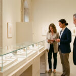 Vue intérieure d’une exposition parisienne de bijoux contemporains, avec des vitrines épurées présentant des broches et des bagues sculpturales, tandis que deux femmes et un homme observent les pièces sous une lumière douce.
